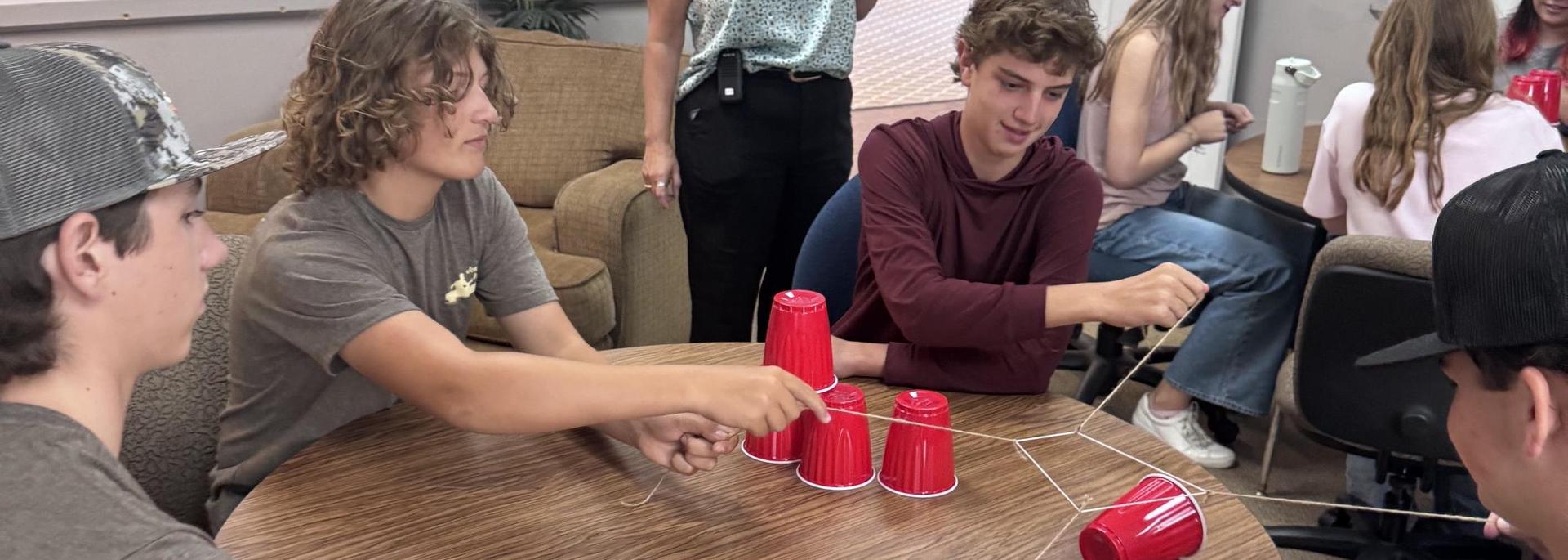 Group of teenagers playing a game with red cups and string at a table.