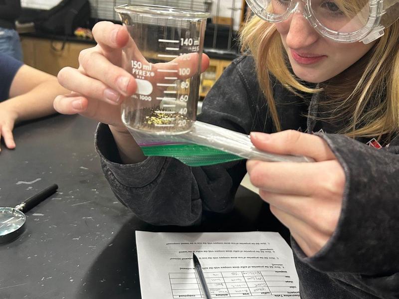 Girl in goggles pouring liquid from a beaker into a bag during a science experiment.