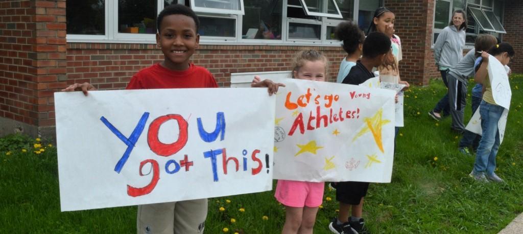 two students hold homemade signs saying "you got this" and "let's go, young athletes"
