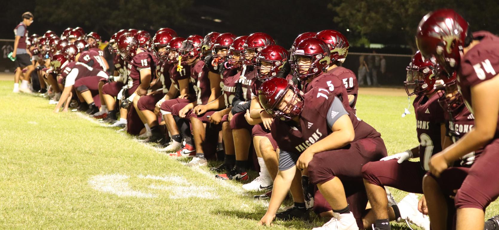 Football team seated on the sideline wearing maroon uniforms at night.