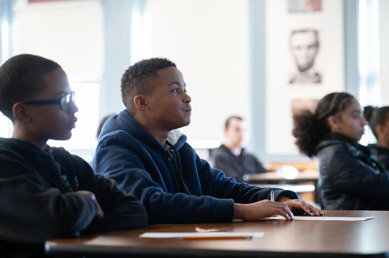 Promise students sit at their desks with light pouring in from the windows behind them