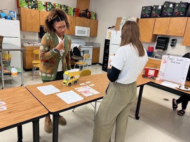 Two individuals discussing at a table in a classroom with food truck displays.
