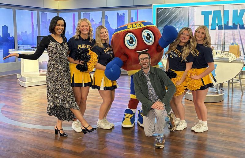 Group of six people, including a mascot, posing with smiles in a studio setting.