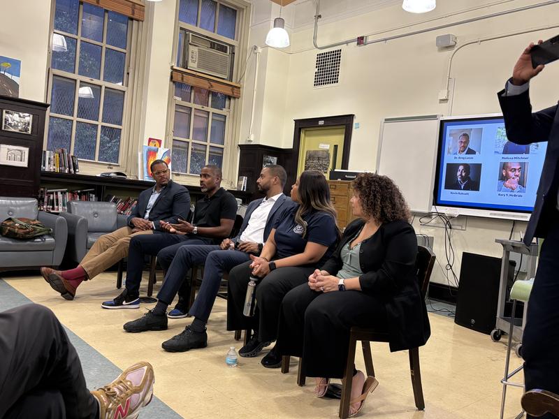 A panel discussion with five speakers seated in a library setting.