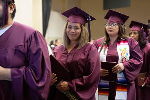 Graduate in maroon cap and gown holding flowers and a diploma smiles gently as she walks in line during the Porterville Adult School graduation.