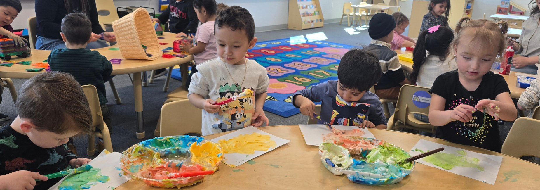Young children engage in art activities at a table covered in paints and drawings.