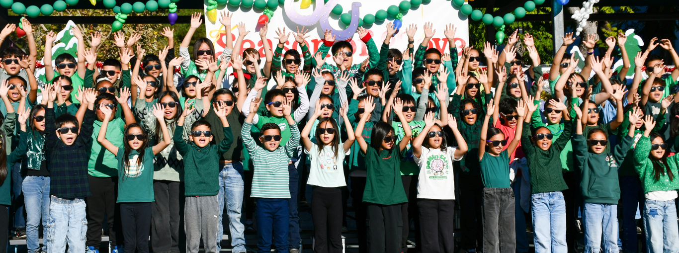 Crowd of children in green outfits joyfully raising their arms in celebration with decorations.