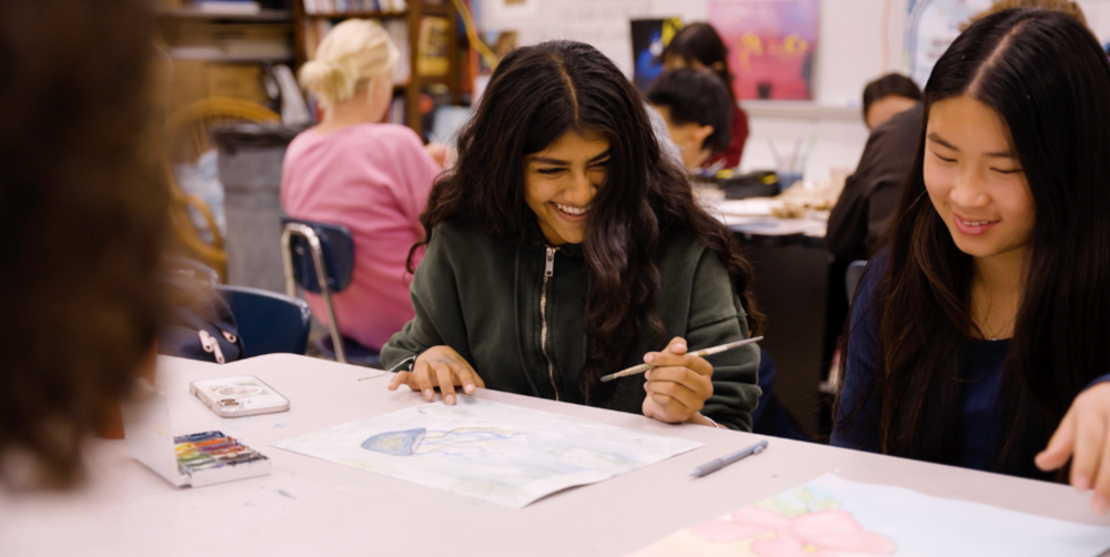 Students engaged in art activities in a classroom, with one smiling as she paints.