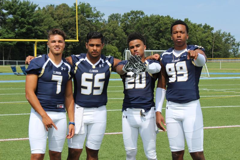 Four football players wearing navy blue jerseys with white and gold trim, white pants, and the word "Clarion" across their chests stand on a grass field with a goalpost in the background.