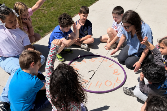 Group of children and adults sitting in a circle, engaging in an outdoor activity.