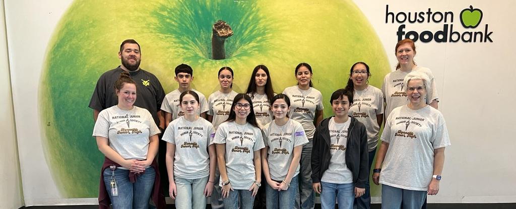 Group of students and teachers posing in front of a large green apple mural.