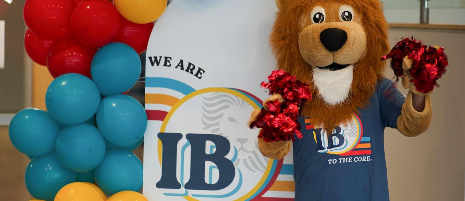 Mascot Roary holds red pom poms in front of a sign with balloons saying, "We are IB to the Core"