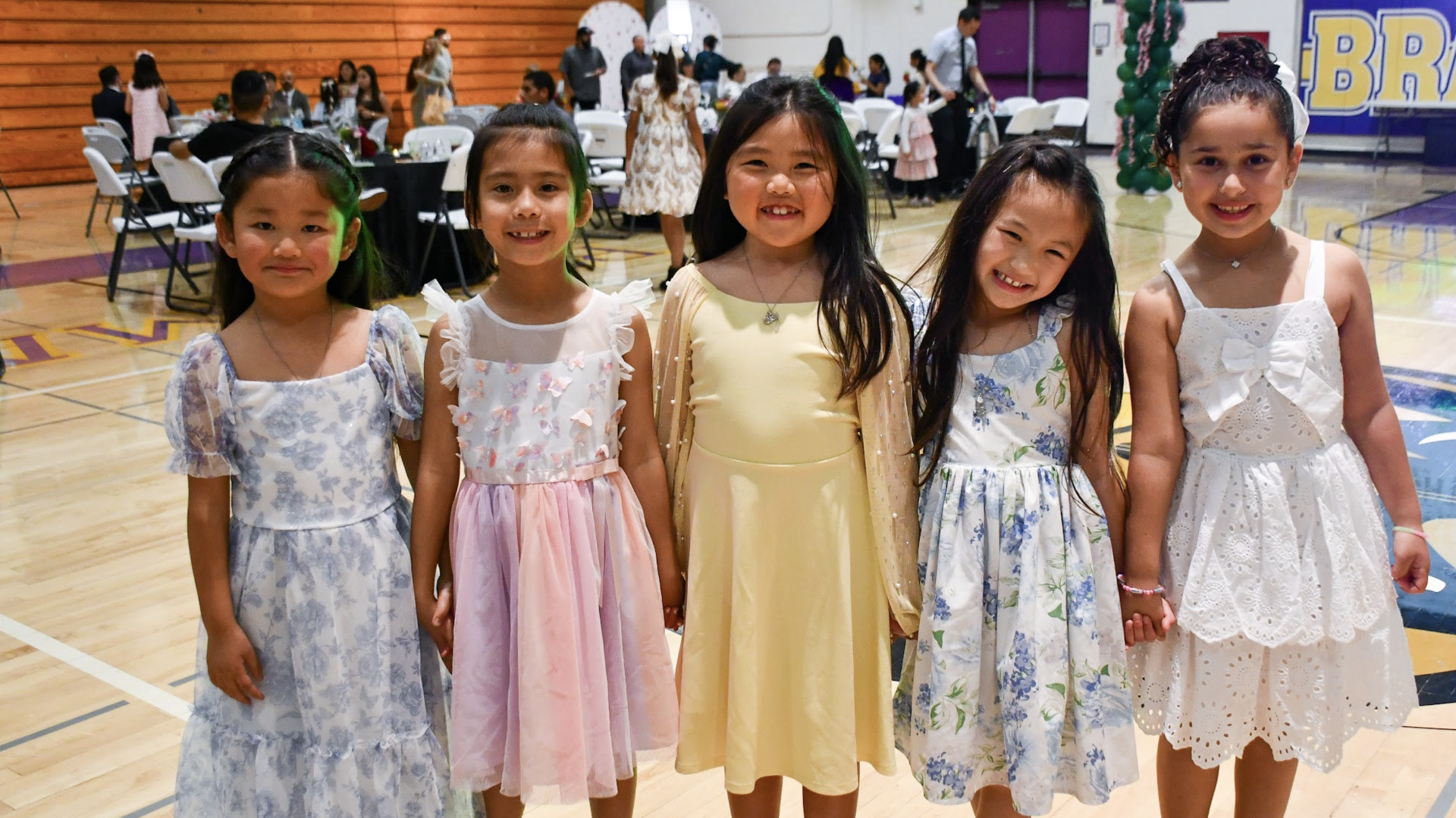Five girls in colorful dresses smiling together at a lively event.