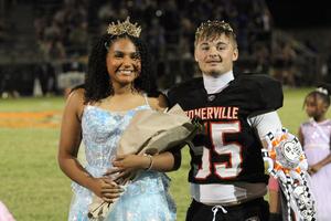 A smiling girl in a blue dress stands next to a football player with a crown.