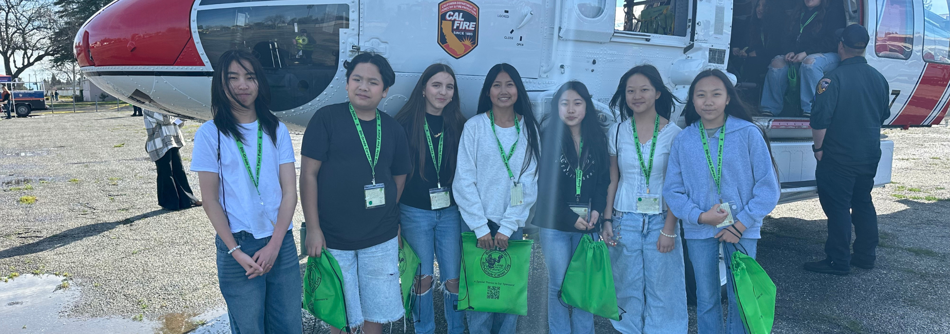 Eight students pose with green bags near a helicopter on a sunny day.
