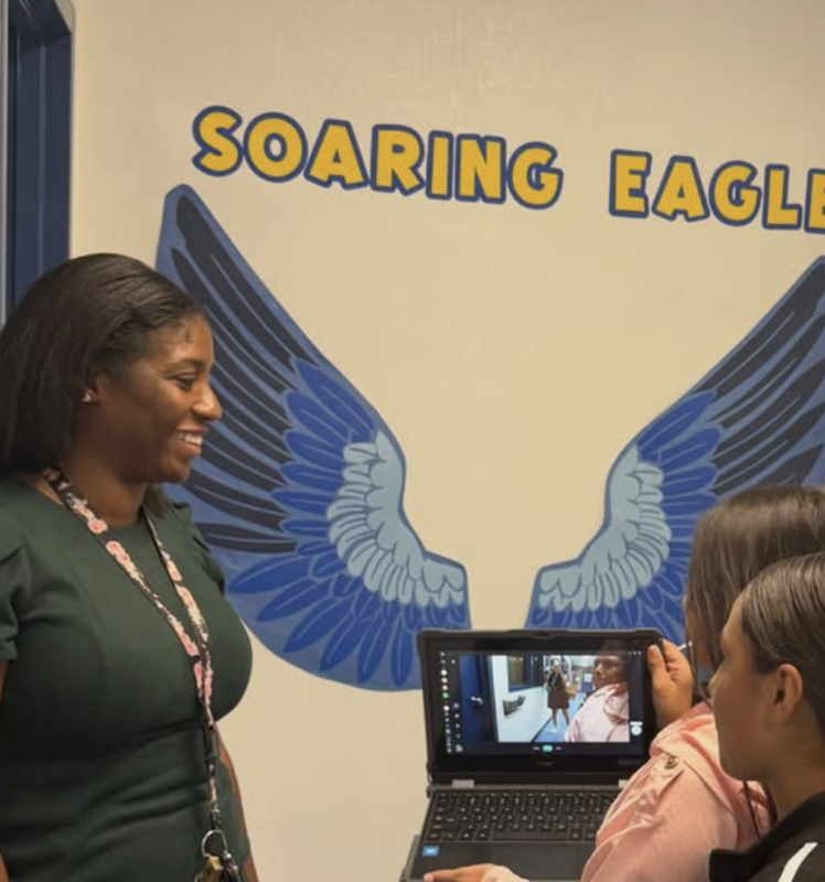 students hold up laptop to show screen to teacher