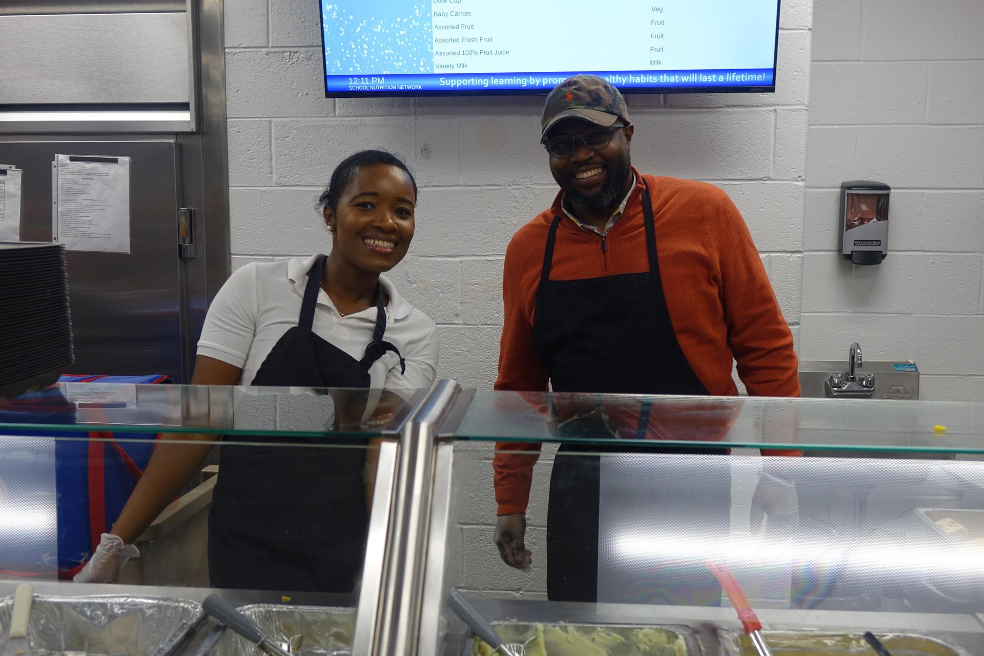 Smiling staff members behind a serving counter in a cafeteria.