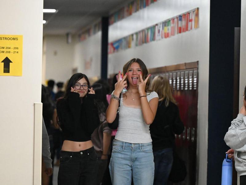 Two students posing playfully in a school hallway with other students in background.