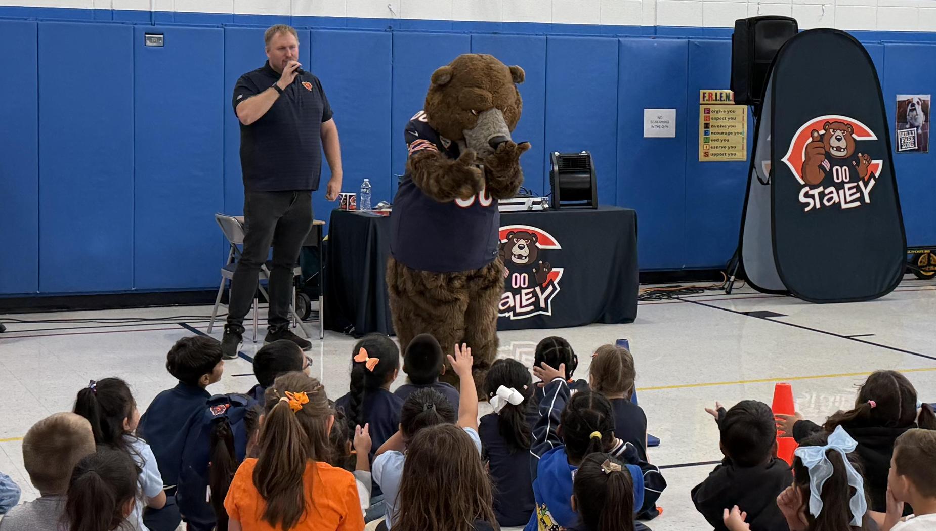 A bear mascot entertains children while a man speaks at an event.