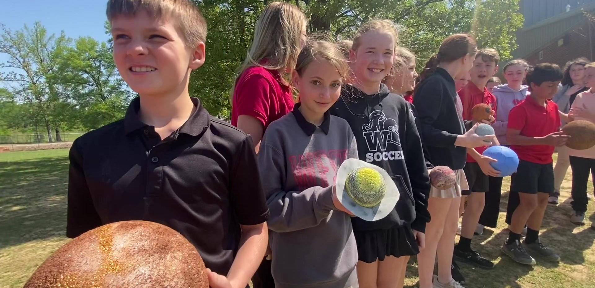 Students in line, smiling and holding various colorful balls outdoors.