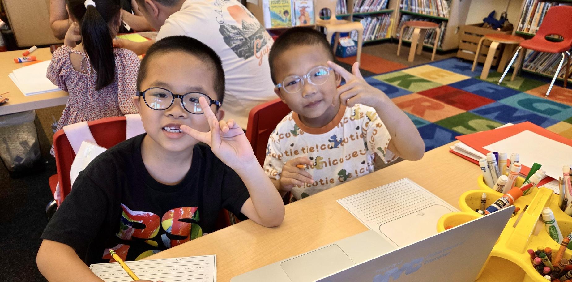 Two children sitting at a table with school supplies, making peace signs.