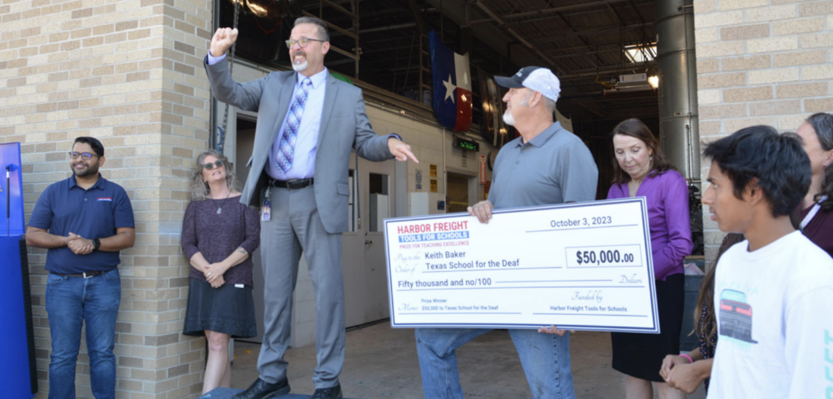 Superintendent Peter Bailey, standing on a ledge and gesturing enthusiastically, presents an oversized check for $50,000 to Keith Baker of the Texas School for the Deaf. The check, dated October 3, 2023, is from Harbor Freight Tools for Schools as part of their Prize for Teaching Excellence. Surrounding them are smiling onlookers, including school staff and community members, celebrating the achievement outside the school's workshop area. A large Texas flag is visible in the background, symbolizing the school’s pride and local support.
