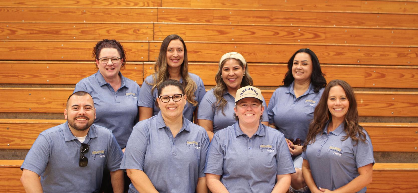 Group of women in blue shirts smiling while seated on wooden bleachers.
