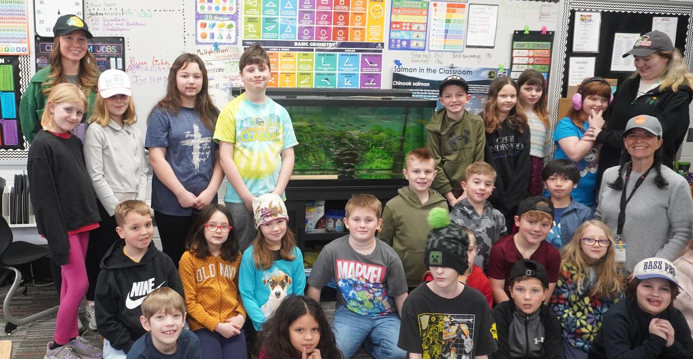 A Lee class poses in front of the fish tank filled with tiny salmon.