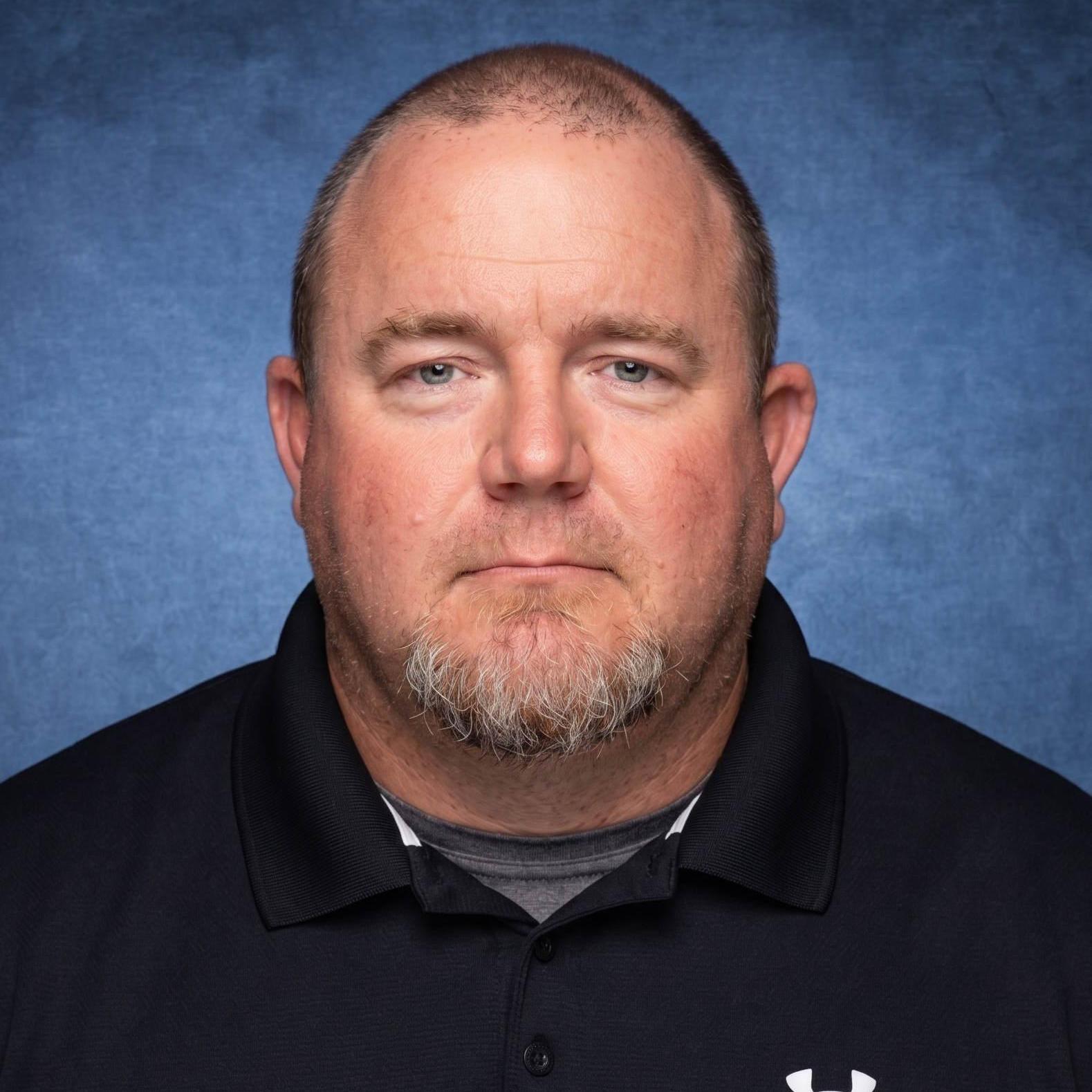 Man with short hair and beard, wearing a black polo shirt against a blue background.