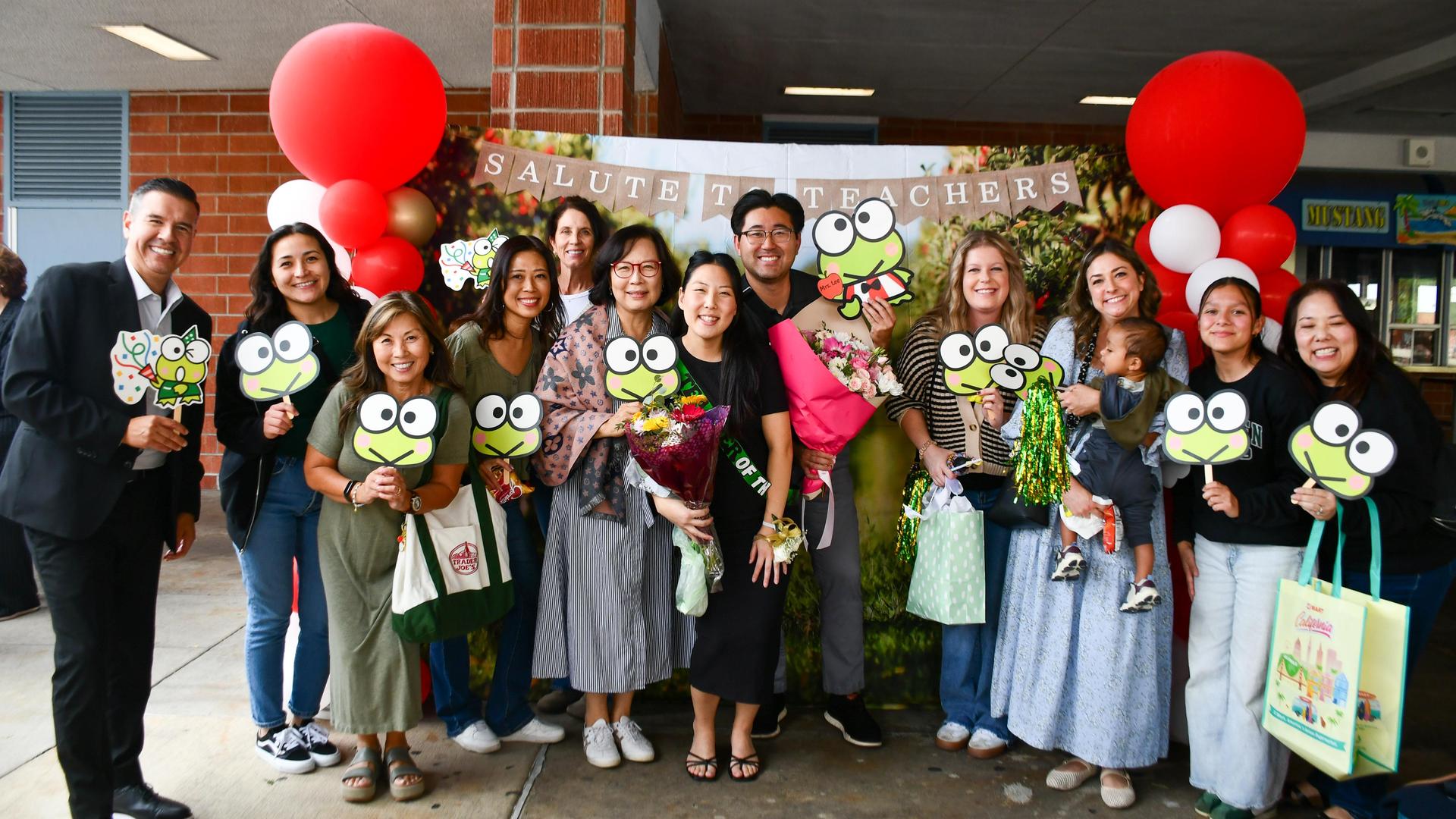 Group of people celebrating teachers with frog-themed signs and balloons.