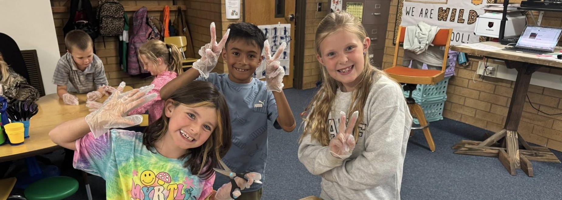 Three children smiling and showing peace signs while working at a table.