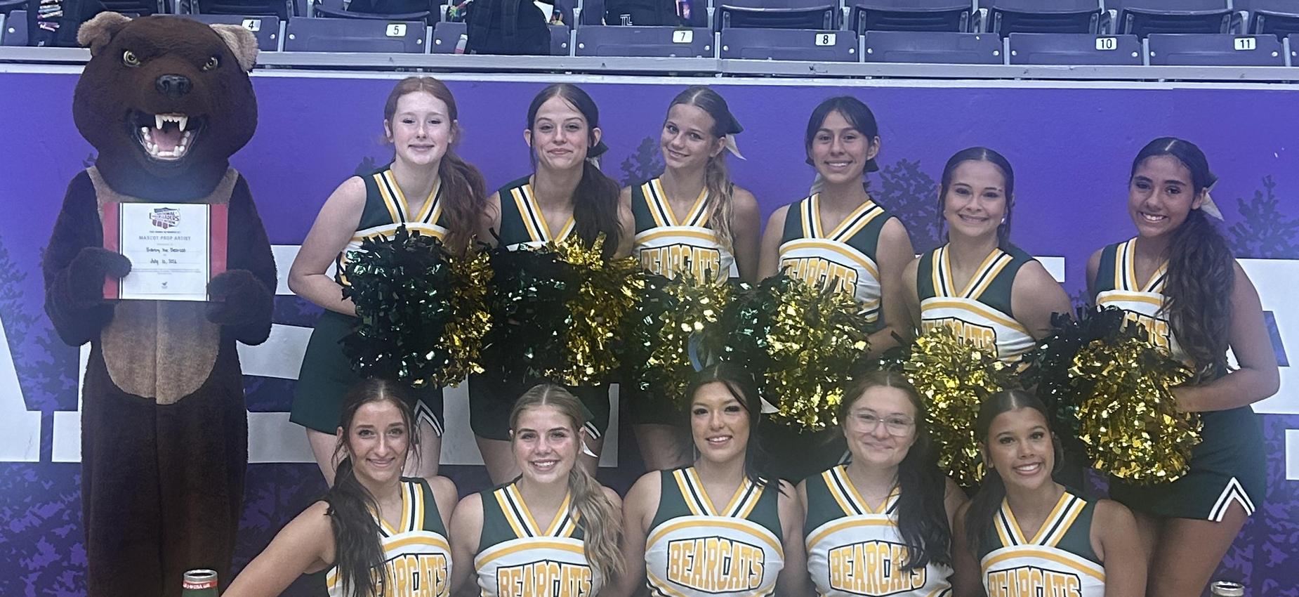 Cheerleading squad posing with a bear mascot and banners at an event.