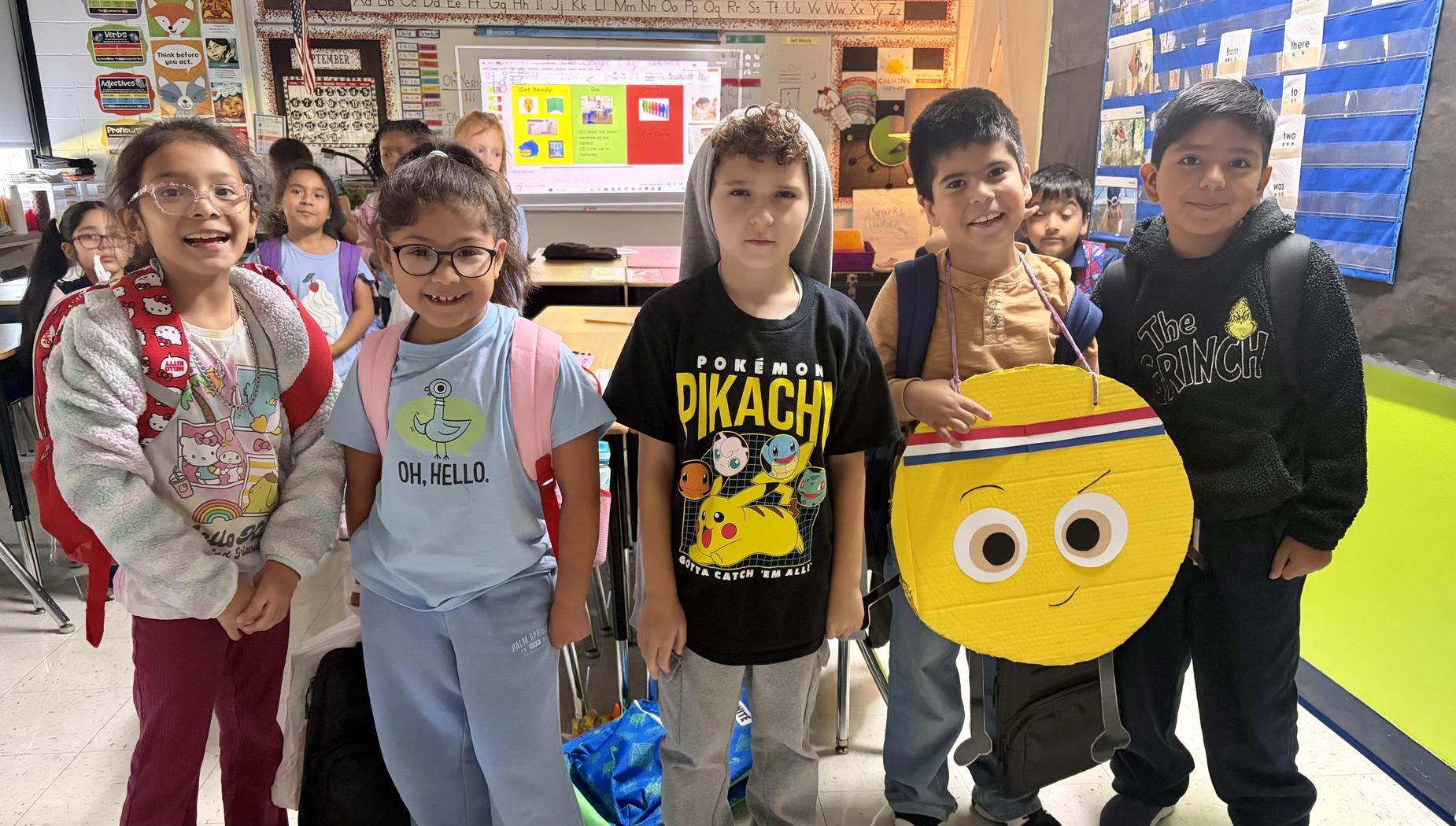 Group of children posing together in a classroom, smiling and wearing backpacks.