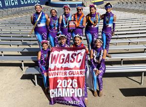 A group of color guard students smile at the camera while holding up their championship banner.
