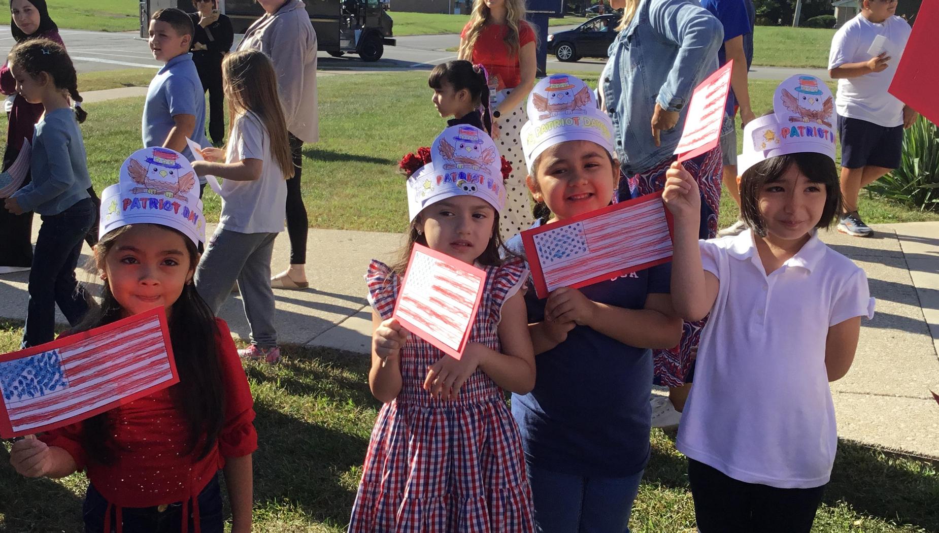 Four children holding American flags, wearing decorated hats, outdoors at an event.