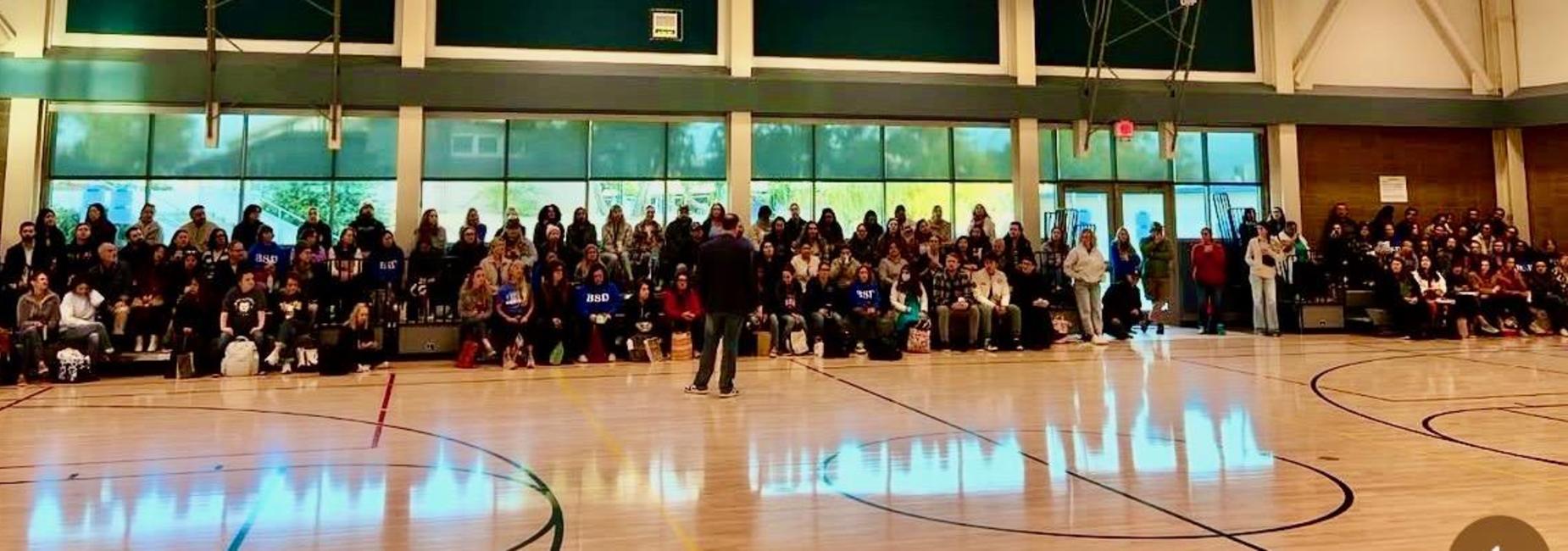 Large crowd seated indoors, viewing a speaker on a gym floor with bright windows.