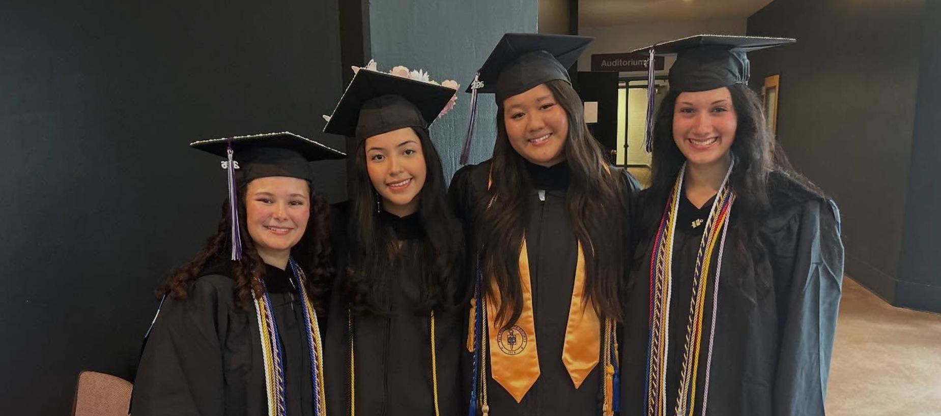 Four graduates in caps and gowns posing together with smiles.