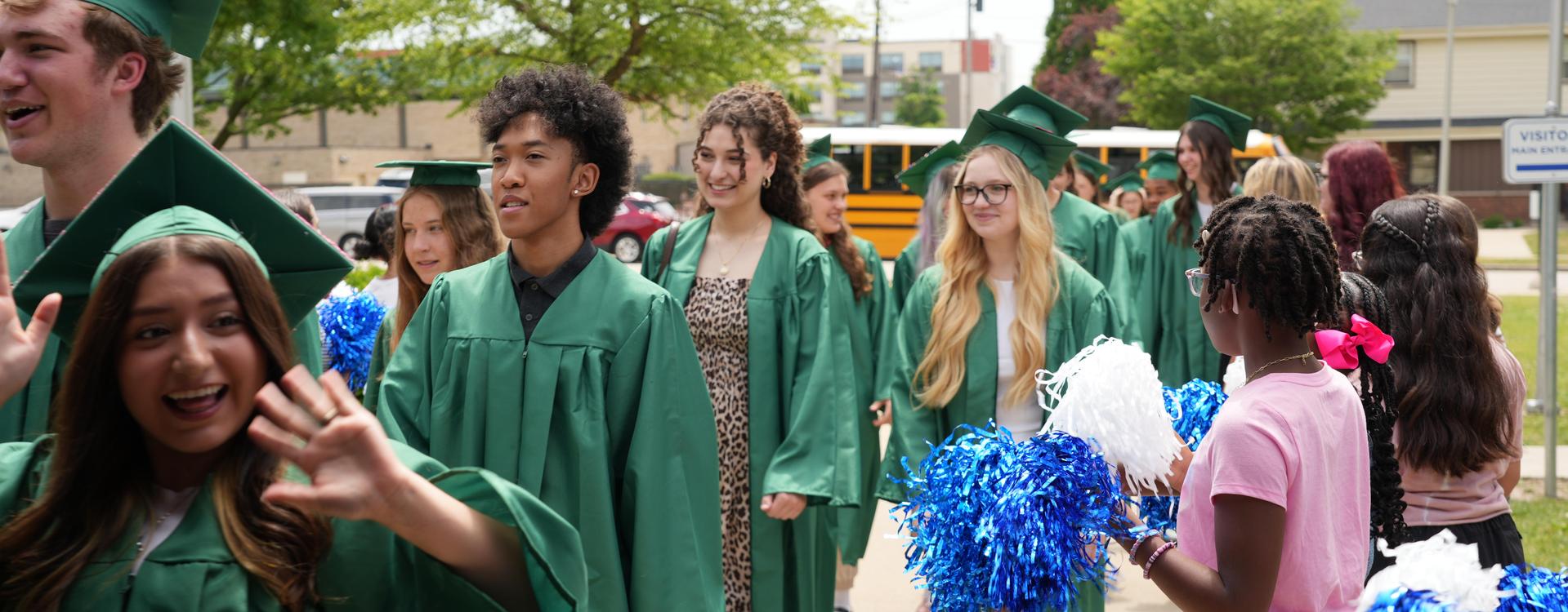 A group of graduates in green gowns walks, while some cheer with pom-poms.