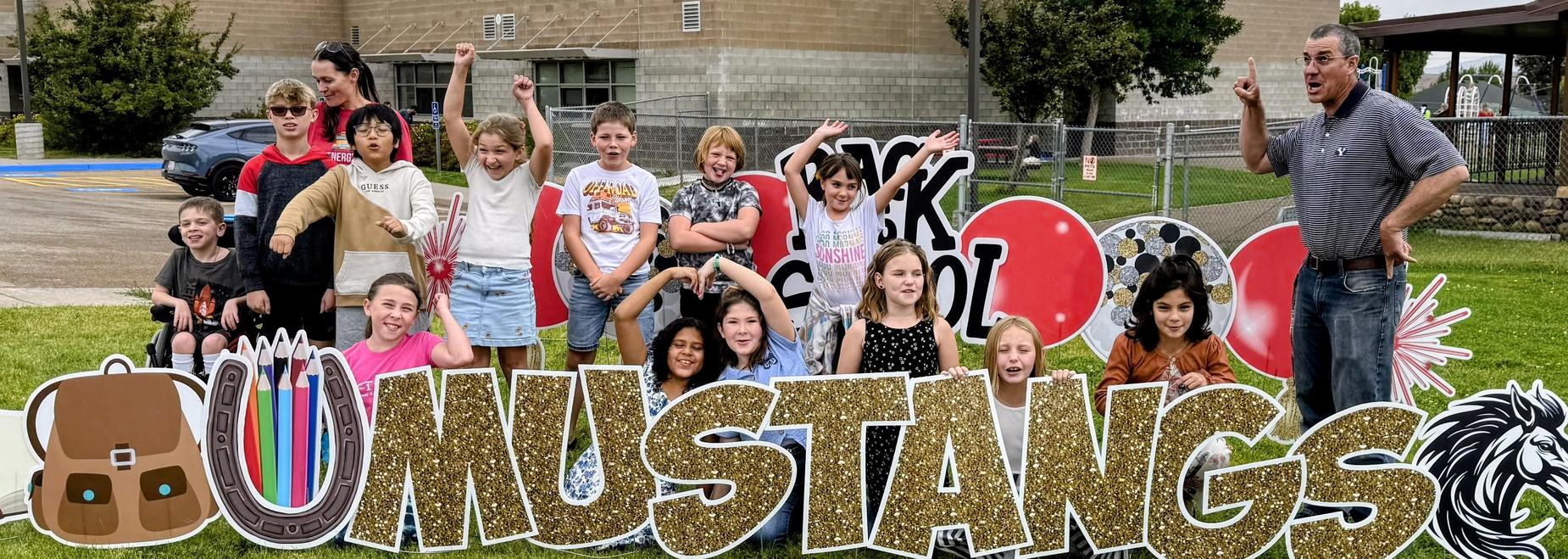 Group of children celebrating with a large Mustangs sign in a grassy area.