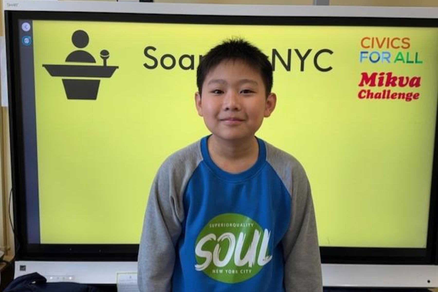 A young boy stands in front of a bright yellow screen with logos and text.