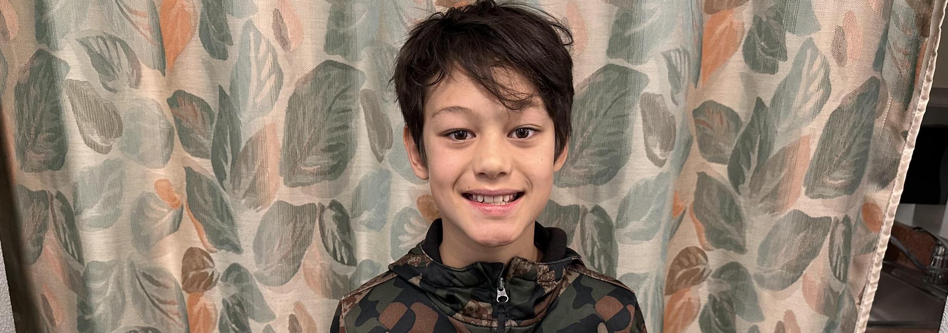 Smiling boy in a camouflage jacket stands in front of a leafy curtain.