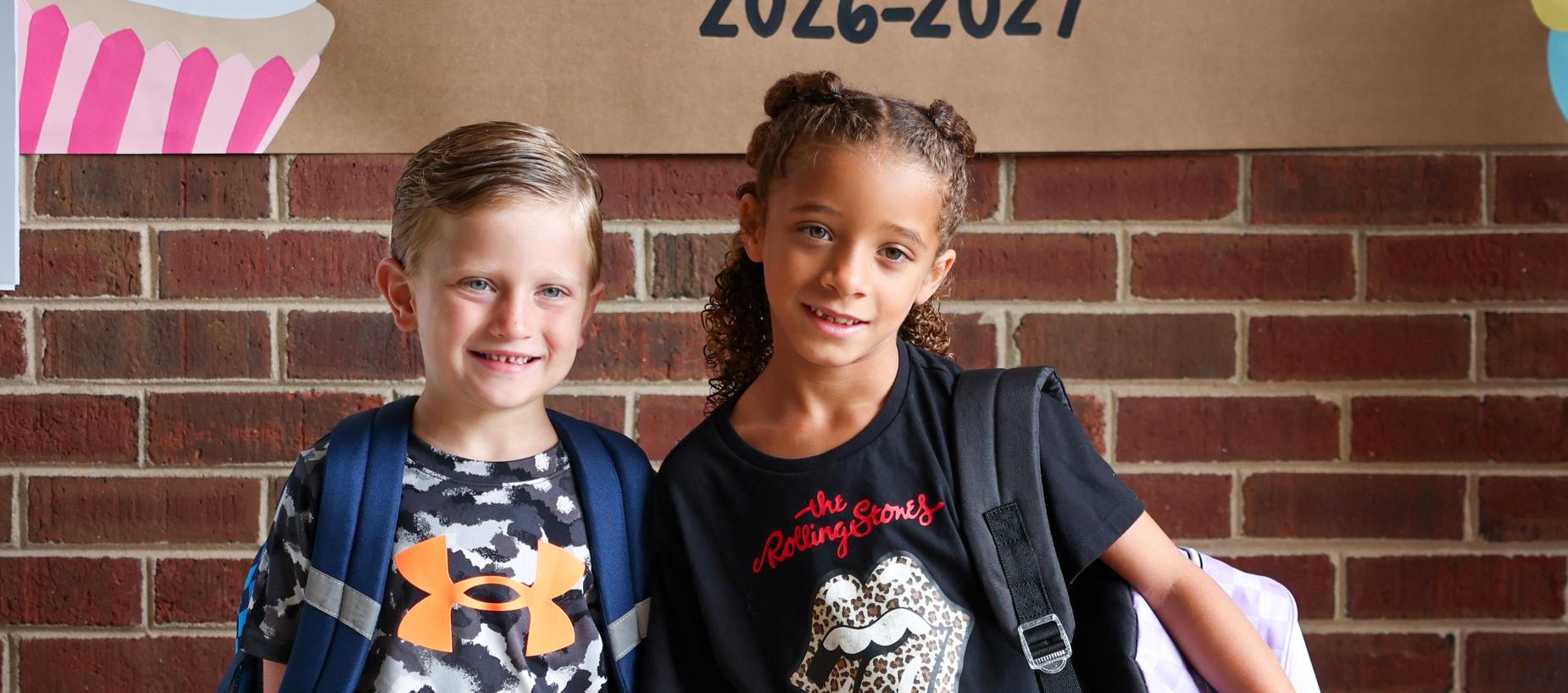 Two children smiling together in front of a brick wall, each with backpacks.
