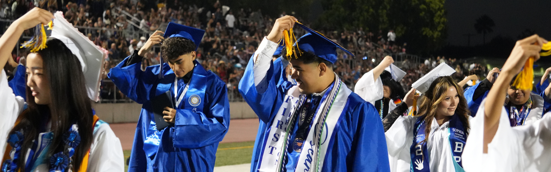 Graduates in blue caps and gowns adjust their tassels during a night graduation ceremony.