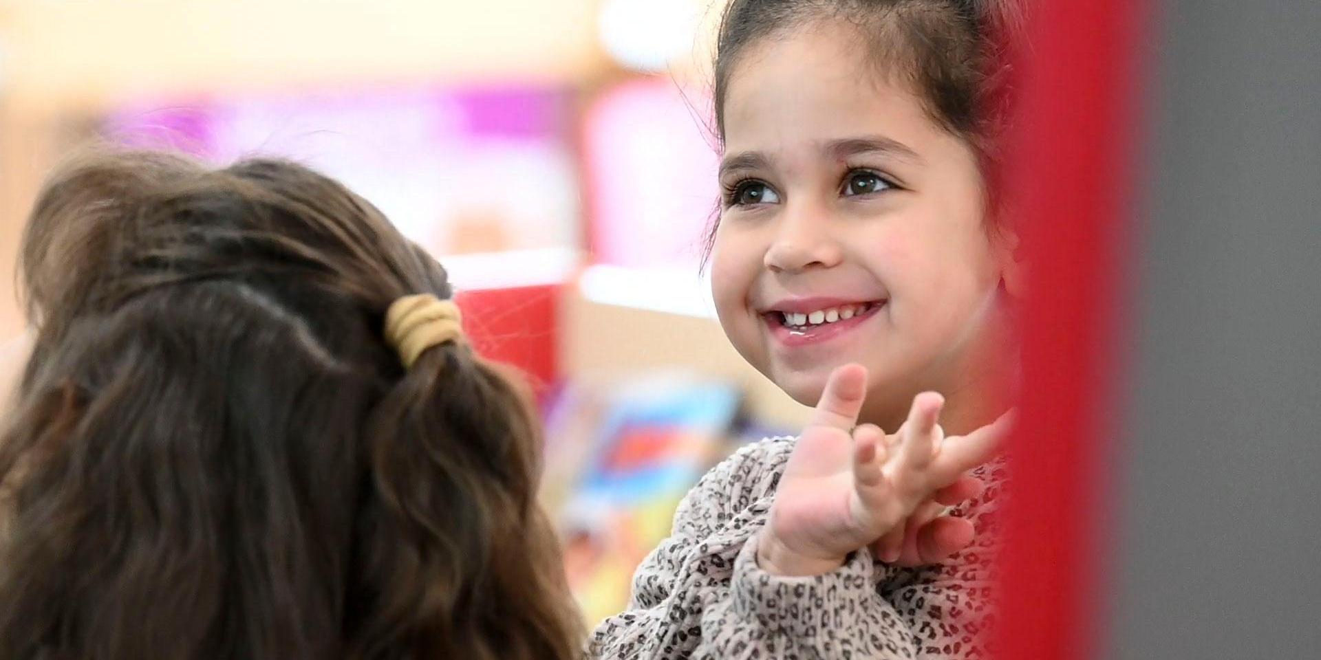 A smiling girl waving, engaged with another child in a colorful indoor setting.