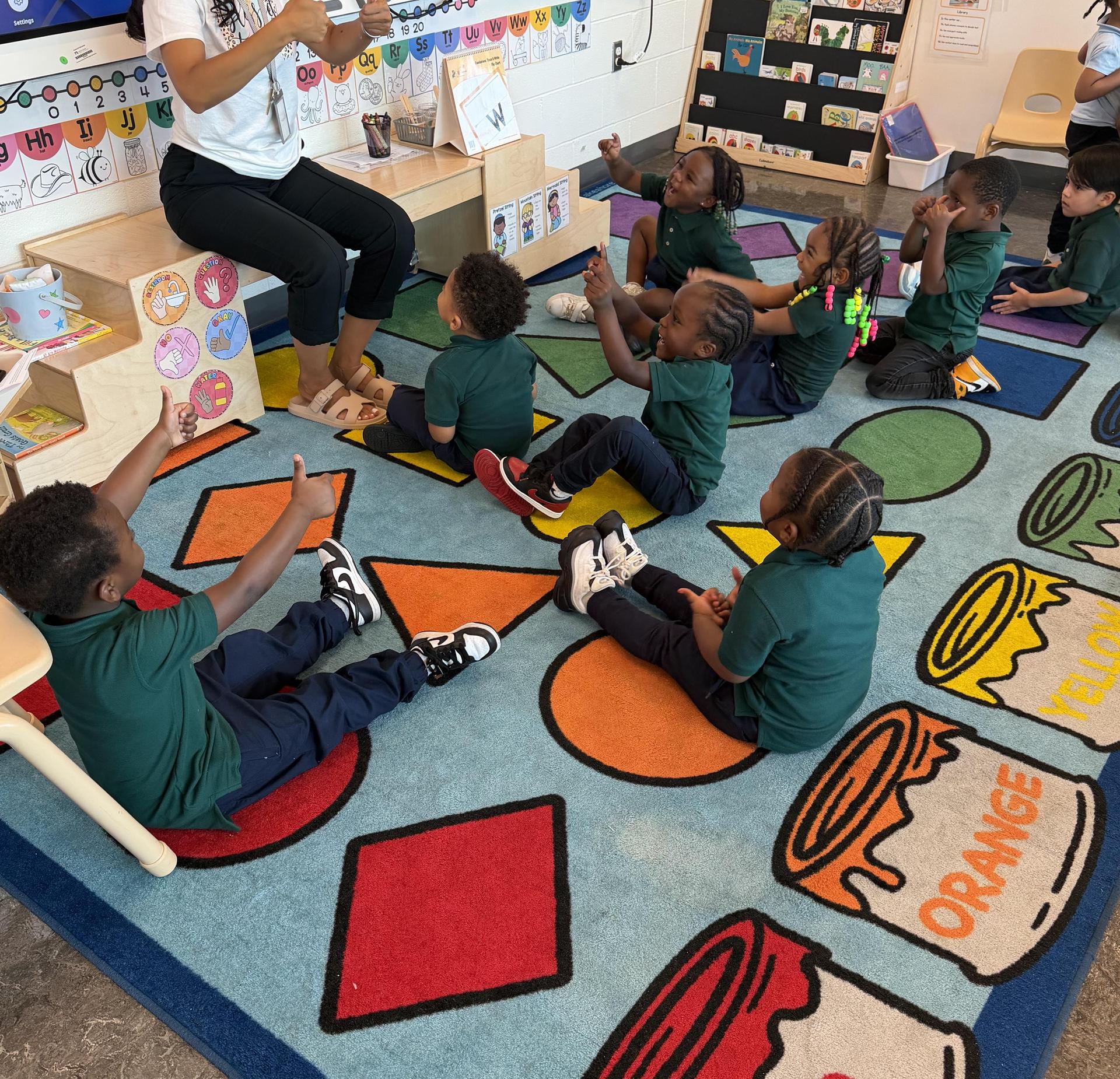 Children sitting on a colorful carpet listening to a teacher's instruction.
