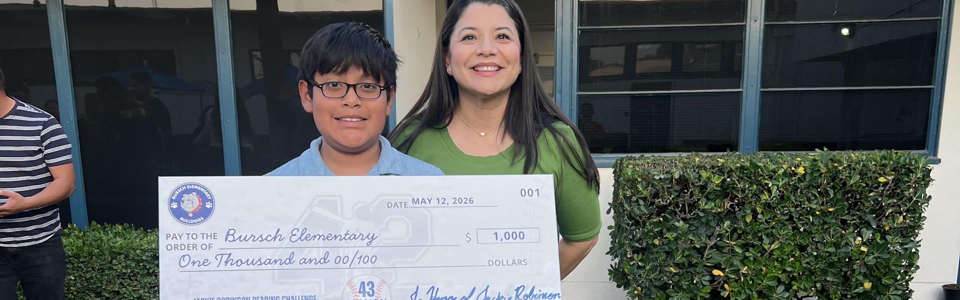 Two individuals hold a large mock check for one thousand dollars outside a school.