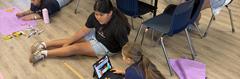 Children sitting on a classroom floor working on projects with laptops and art supplies.