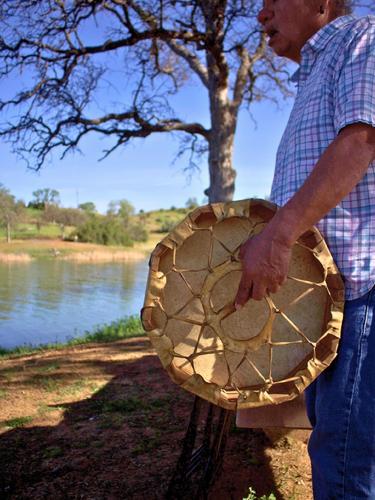 Man holding a drum by a lake with trees and blue sky in the background.