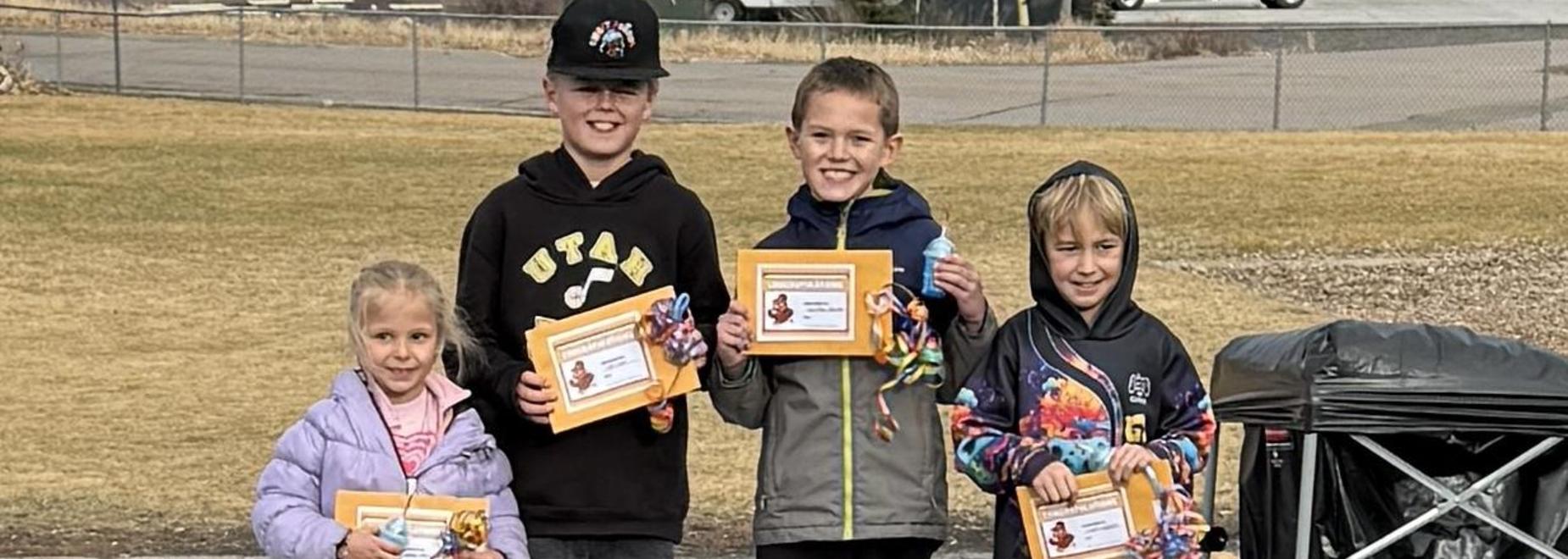 Four children holding certificates and smiling outdoors.