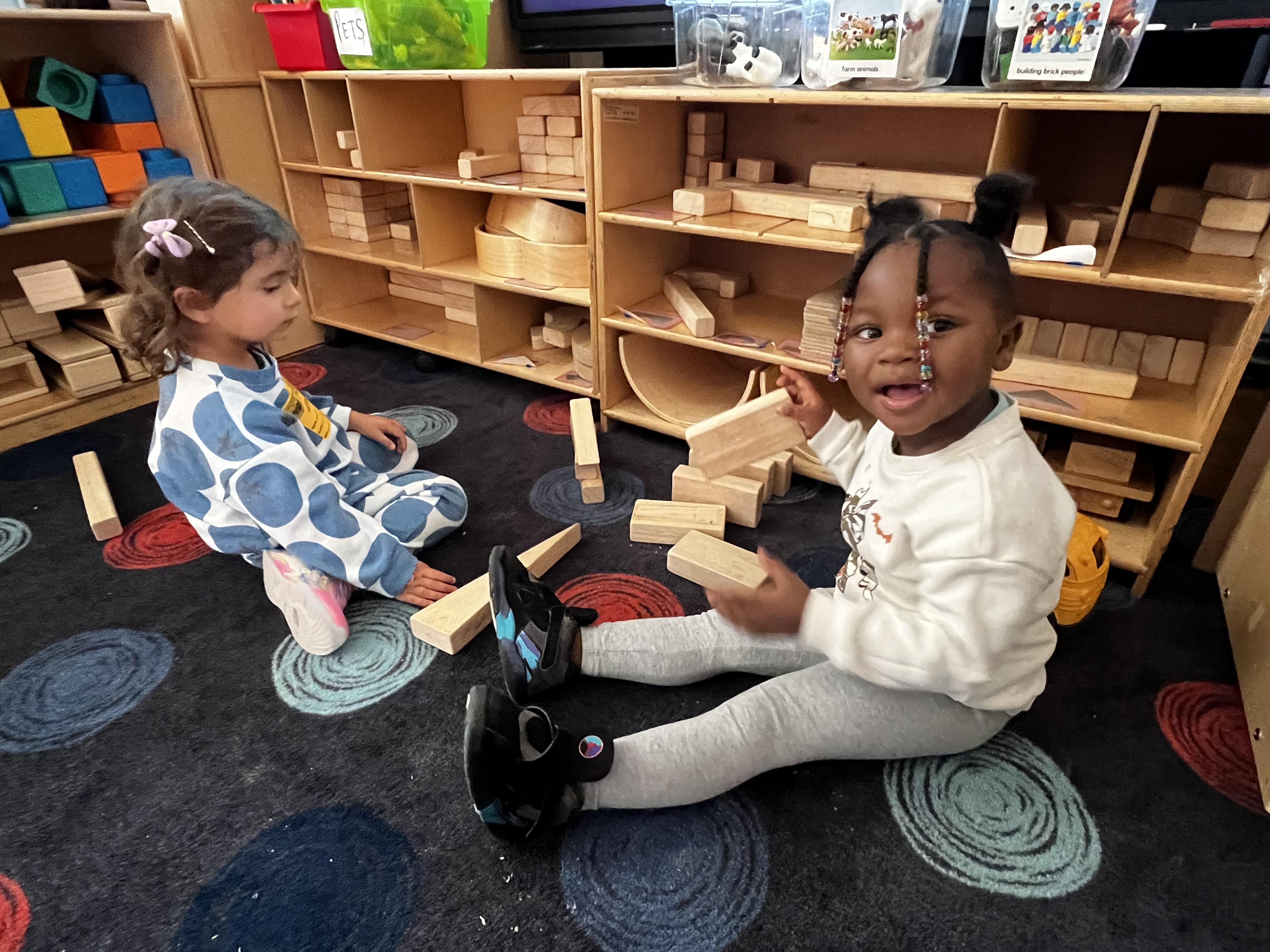 Two young children sit on a patterned rug playing with wooden building blocks next to a shelf full of toys and materials.